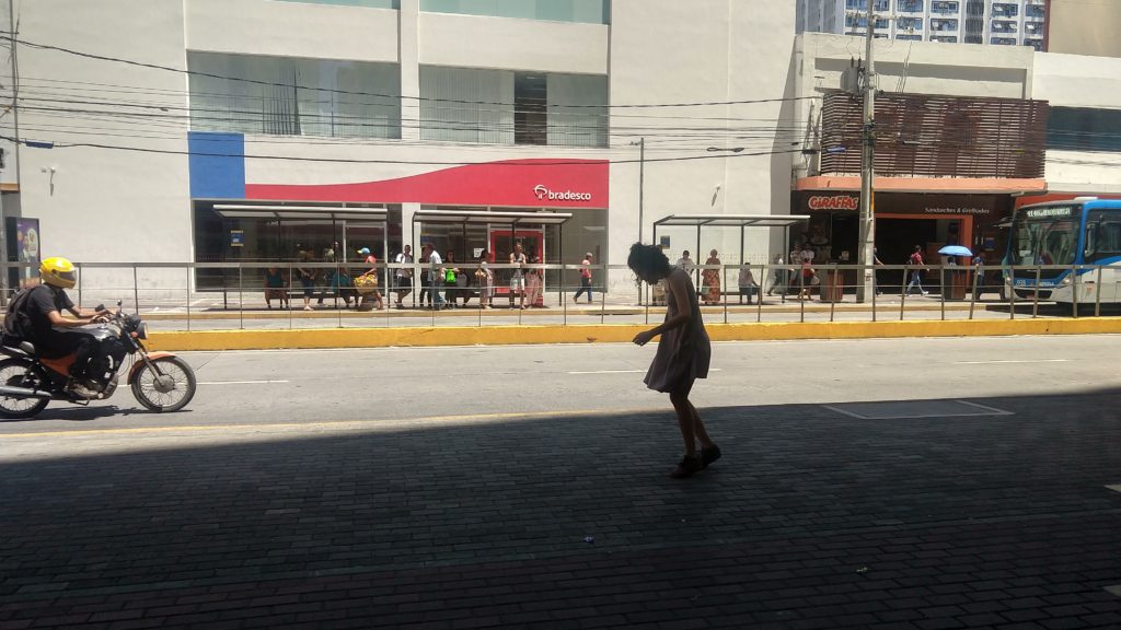 Foto da calçada de uma avenida. Ao fundo, vemos um ponto de ônibus cheio de pessoas e a fachada do Banco Bradesco. No primeiro plano, do outro lado da avenida, Amanda dança, ela tem seu vestido e cabelos esvoaçando. 