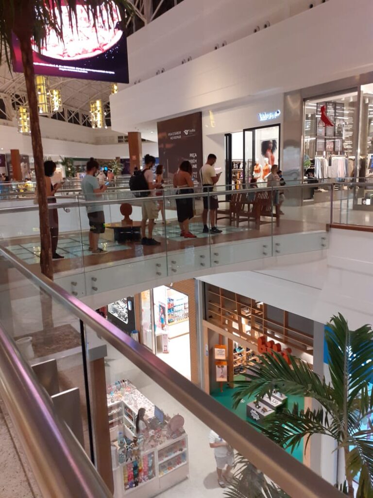 Foto de um shopping. Seis pessoas andam em fila, elas tem a cabeça baixa e o olhar no celular.