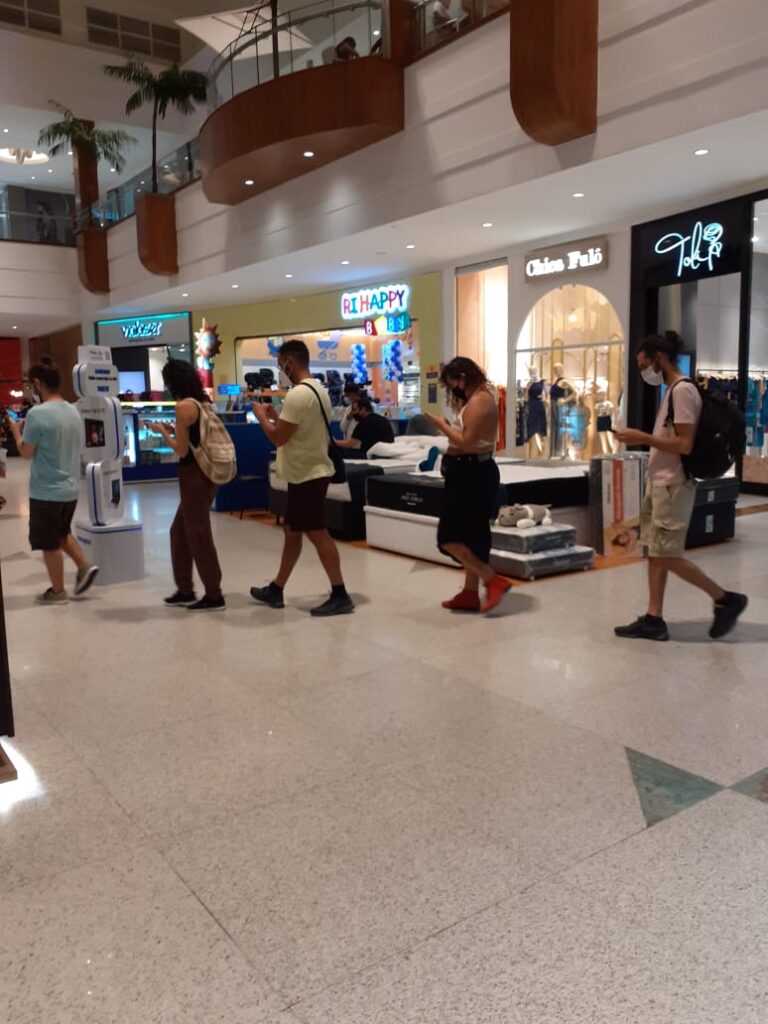Foto de um shopping. Vemos a fila de pessoas andando, uma vendedora de um kiosque do shopping ignora a fila e segue olhando para seu computador.